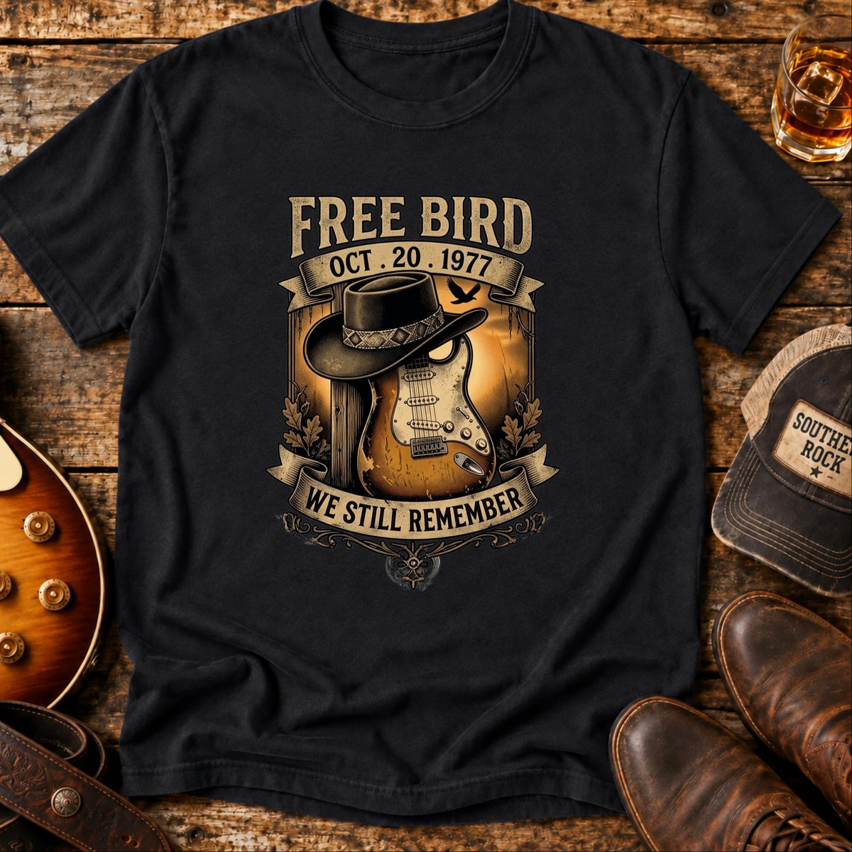 We Still Remember tribute tee in black — Free Bird artwork with a black wide-brim hat resting on a sunburst Stratocaster and the date Oct 20 1977, laid on weathered wood with boots, a whiskey glass and a Southern Rock cap
