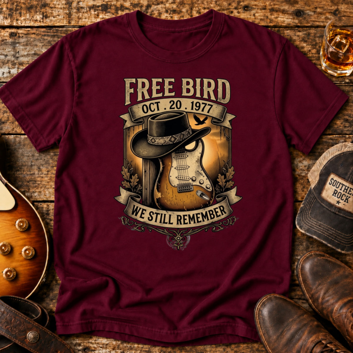 We Still Remember tribute tee in maroon — Free Bird artwork with a black wide-brim hat resting on a sunburst Stratocaster and the date Oct 20 1977, laid on weathered wood with boots, a whiskey glass and a Southern Rock cap